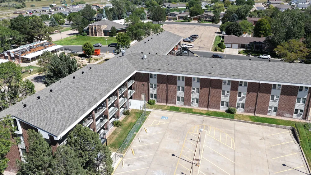 Aerial view of an apartment complex during roofing work, showing a tarped roof section, a completed gray shingle roof, and a boom lift at a balcony.