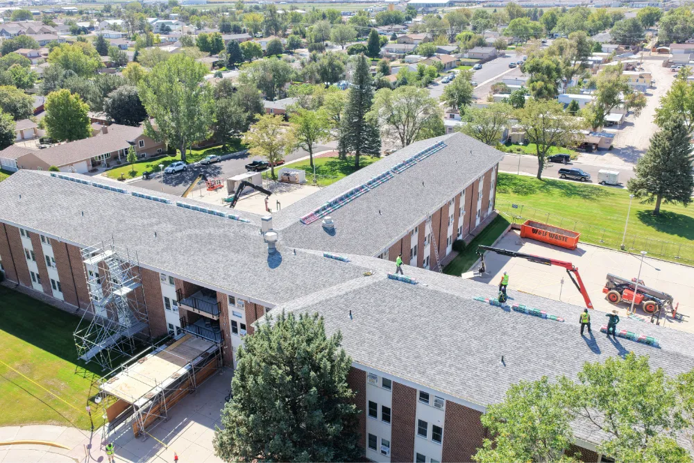Aerial view of an apartment complex during a roof replacement, with scaffolding, a waste dumpster, and crew working on the roof.