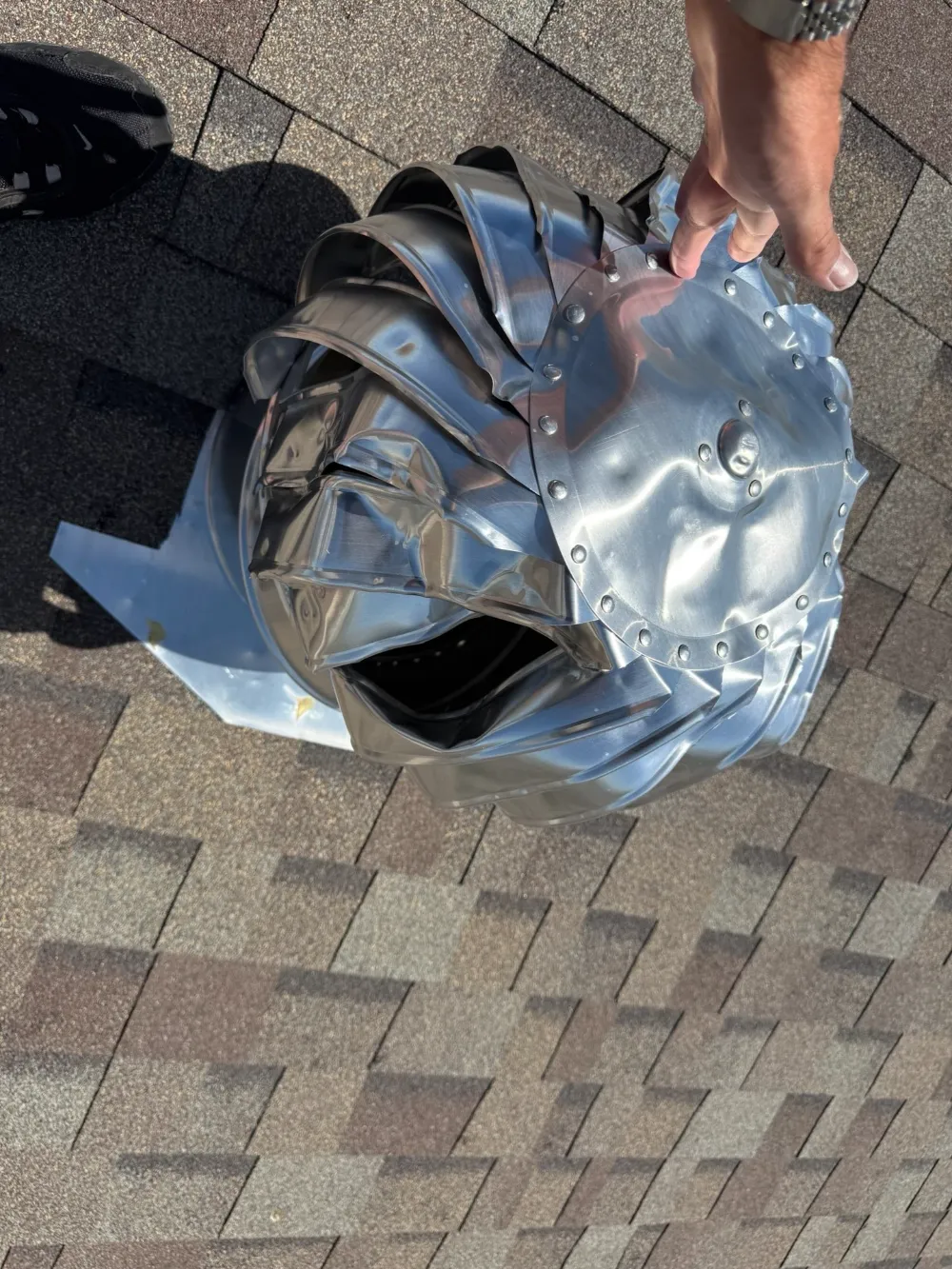 Hand holding a dented and torn metal turbine roof vent removed from a shingle roof.