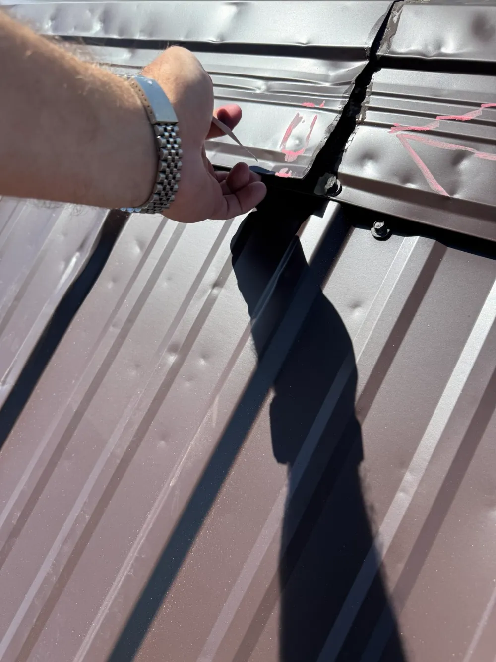 Hand pointing to a split seam in a metal roof panel marked with chalk.