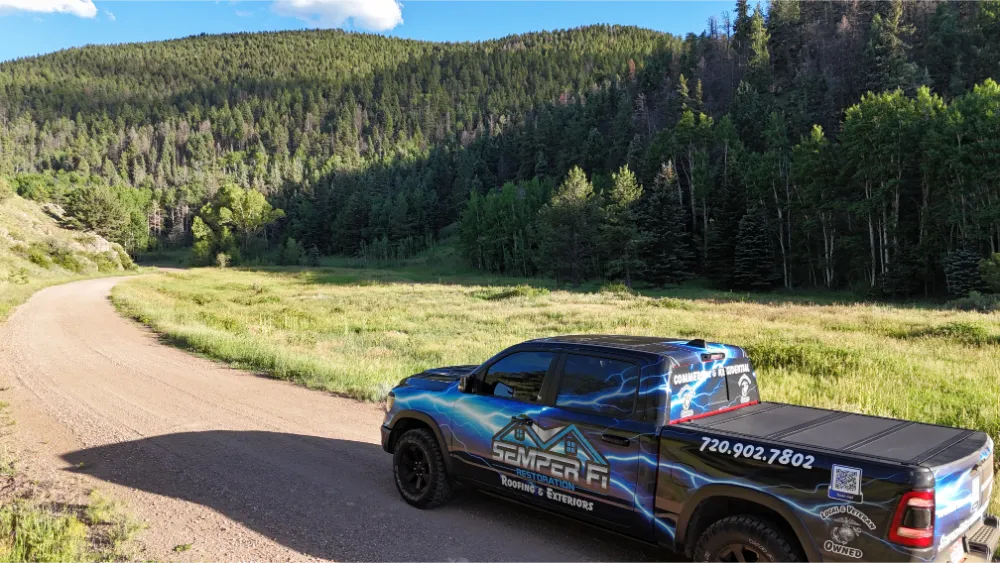 Pickup truck with a vehicle wrap parked on a dirt road in a forested mountain area.