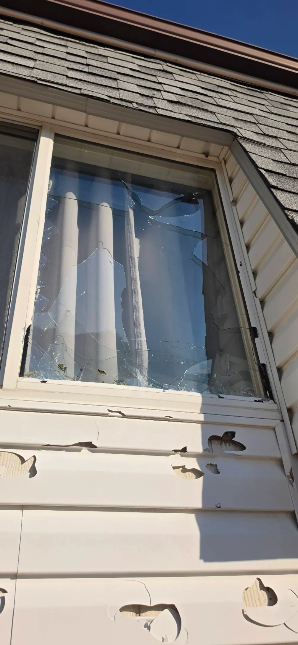 Window with cracked, shattered glass; vinyl siding below shows several impact holes.