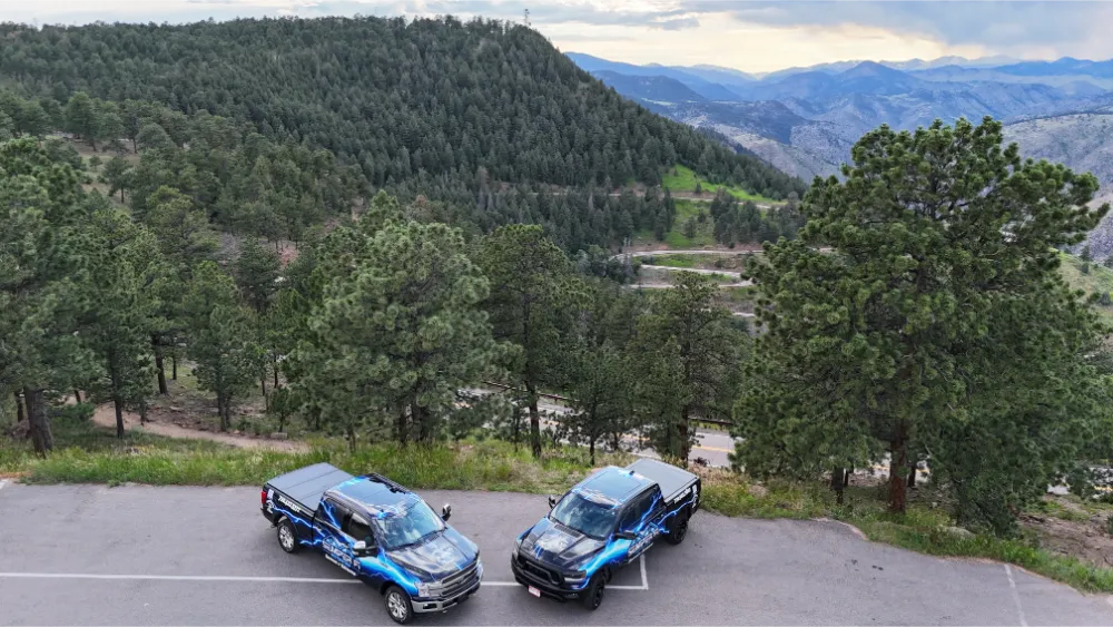 Aerial view of two pickup trucks with matching logo wraps parked at a scenic overlook above a forested valley.